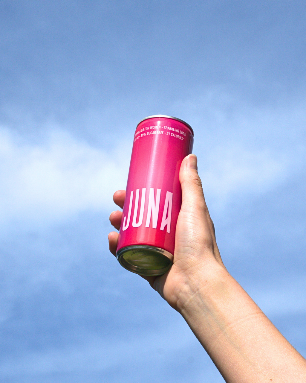 Hand holding a pink JUNA sparkling soda can against a blue sky, representing an on-the-go wellness drink formulated for women